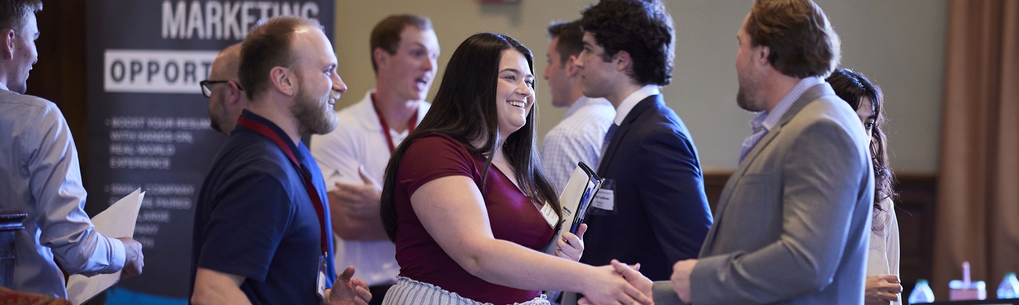 A man and a woman shaking hands at an OU career fair. Many others can be seen in the background talking and networking. 