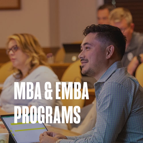 An adult EMBA student sits in class with his peers, listening to the professors lecture. Text over photo reads: MBA & EMBA programs.