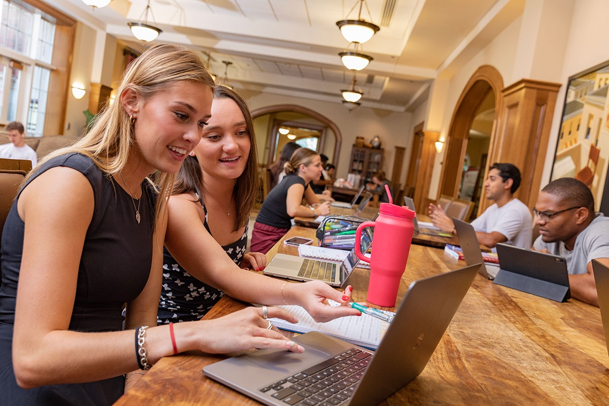 Two students sit together while working at a study table in the Price Hall lounge. Other students are visible in the background. T