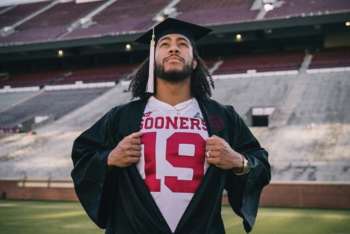 Caleb Kelley stands on the OU Football field, wearing his cap and gown. He is pulling his gown open to display his OU Football Jersey #19
