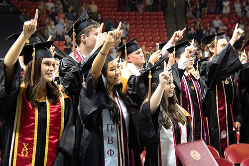 students in cap and gown regalia stand for the OU chant at commencement