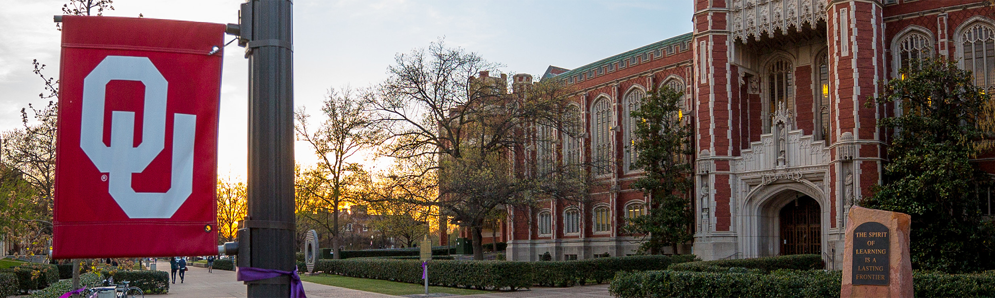 An exterior photo of Bizzell Memorial Library in the morning, with an OU banner handing from a lamppost in the foreground.