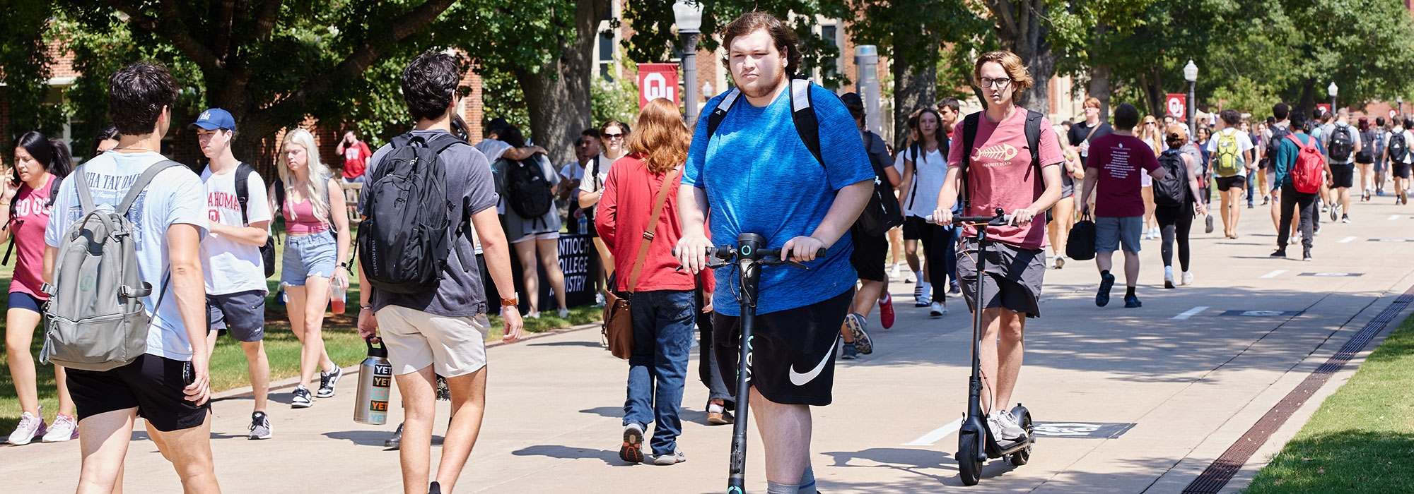 A crowded street on campus with many students walking and riding scooters.