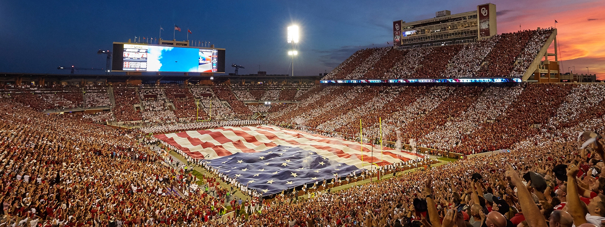 The American flag covering the field at Gaylord Family Oklahoma Memorial Stadium in front of a large crowd.