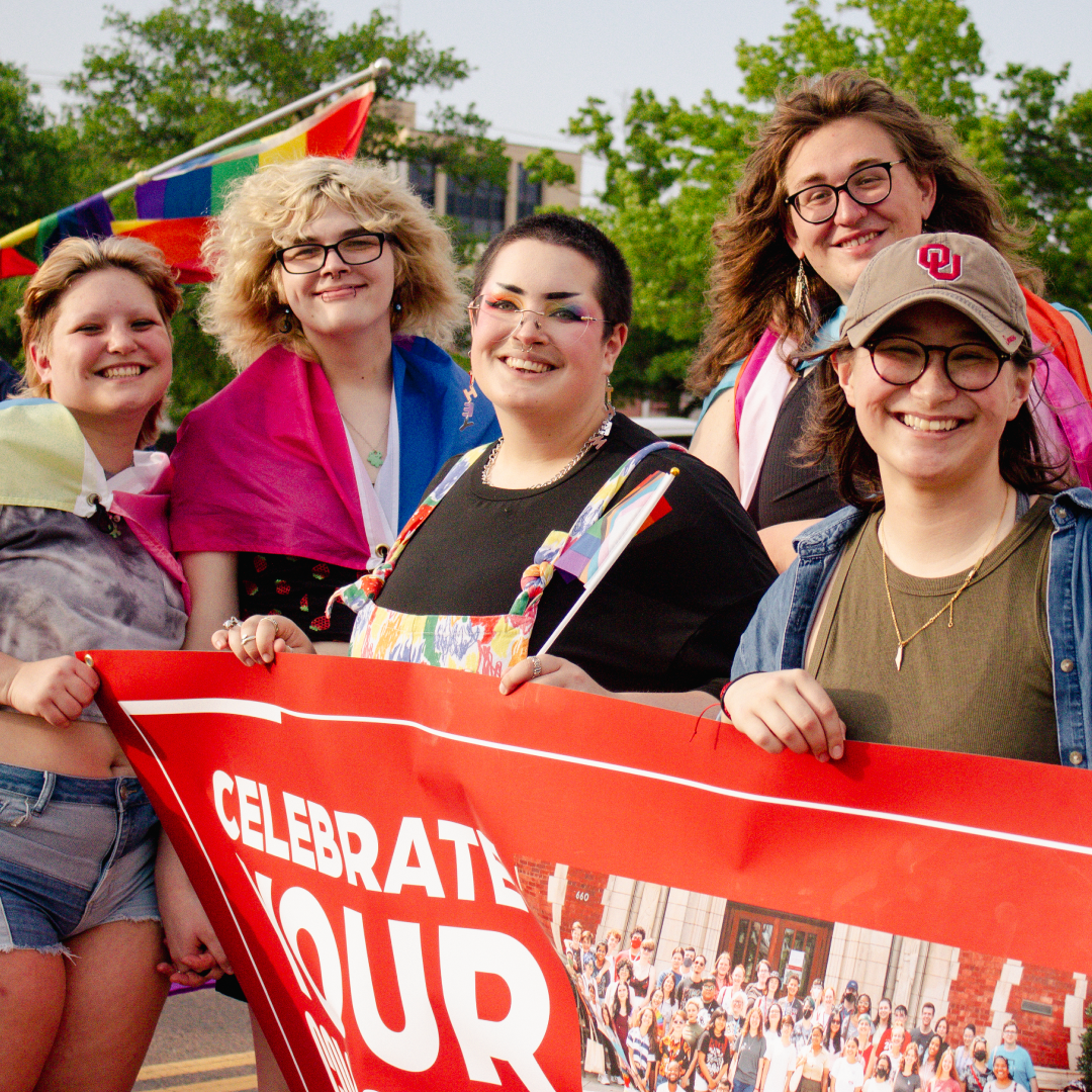 The image shows a group of five smiling individuals standing together outdoors, holding a bright red banner that reads "Celebrate Colors Proudly Gleaming" in bold letters. They are wrapped in colorful pride flags, and a rainbow flag is waving behind them. 