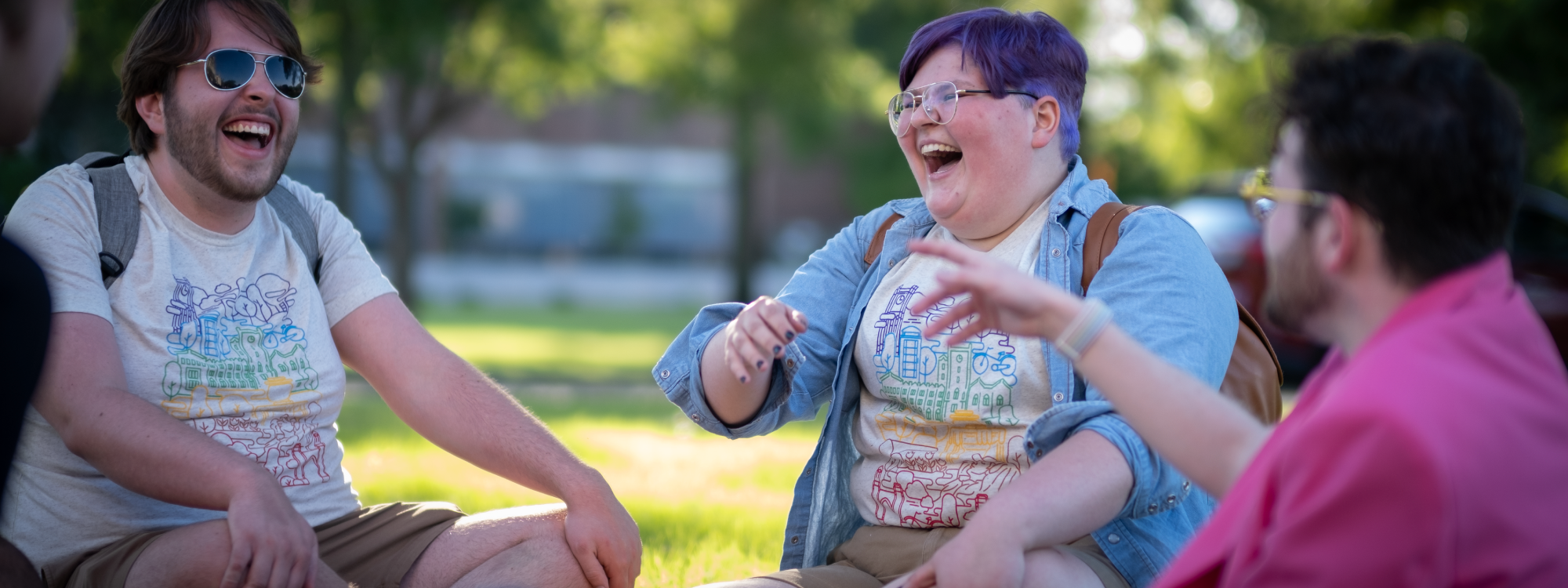 The image shows three people sitting outside on the grass, enjoying each other's company and laughing. They appear relaxed and engaged in a lively conversation, wearing Pride shirts in rainbow. The scene takes place in a sunny, green outdoor setting, creating a cheerful and friendly atmosphere.