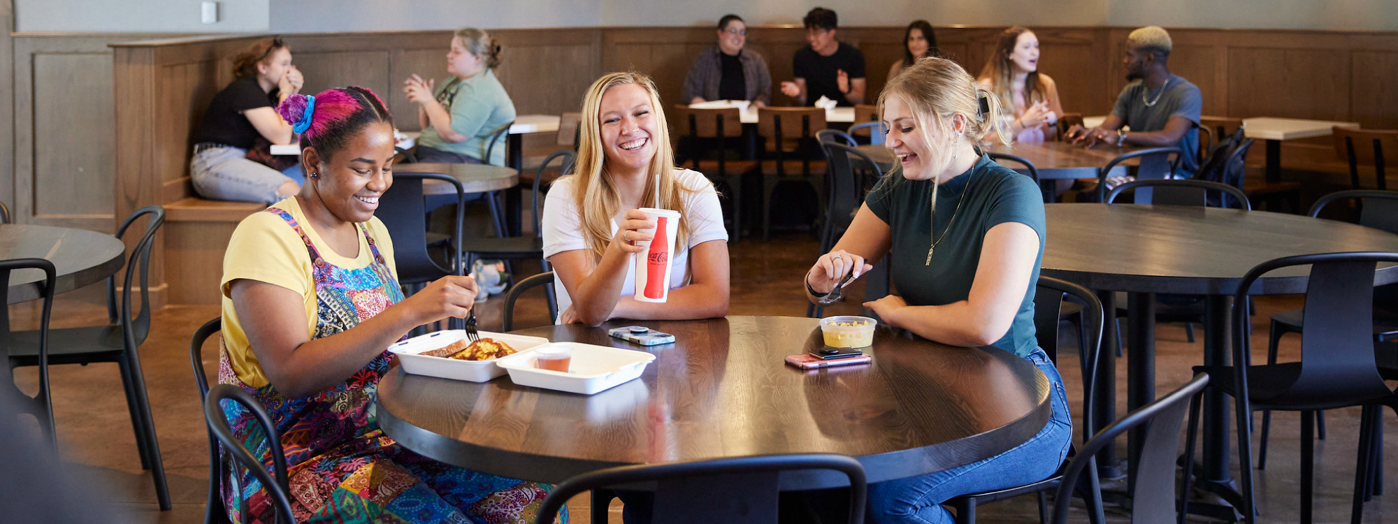 The image shows three young women sitting at a round table in a casual dining area, enjoying their meals and laughing together. In the background, other people are seated at various tables, creating a lively and relaxed atmosphere. The women appear to be engaged in friendly conversation, contributing to the warm and welcoming environment of the space.