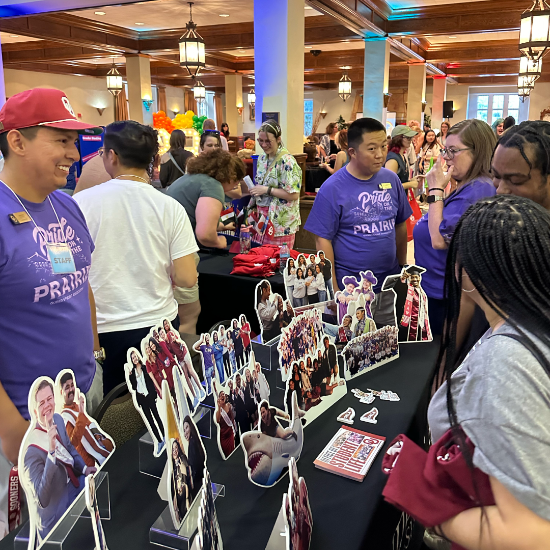 The image shows the LGBTQIA+ Student Welcome where people are engaging with a booth displaying cutouts of individuals in various poses. The booth staff, wearing purple "Pride on the Prarie" shirts, are interacting with visitors in a warm, rainbow-lit space, creating a welcoming and celebratory atmosphere.