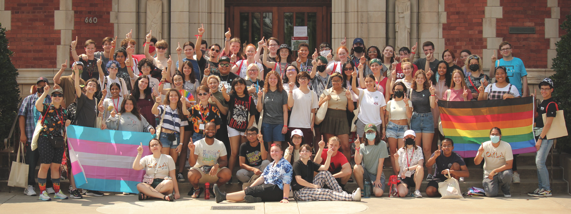 A group of students standing in front of Evans Hall with pride flags holding up their one finger