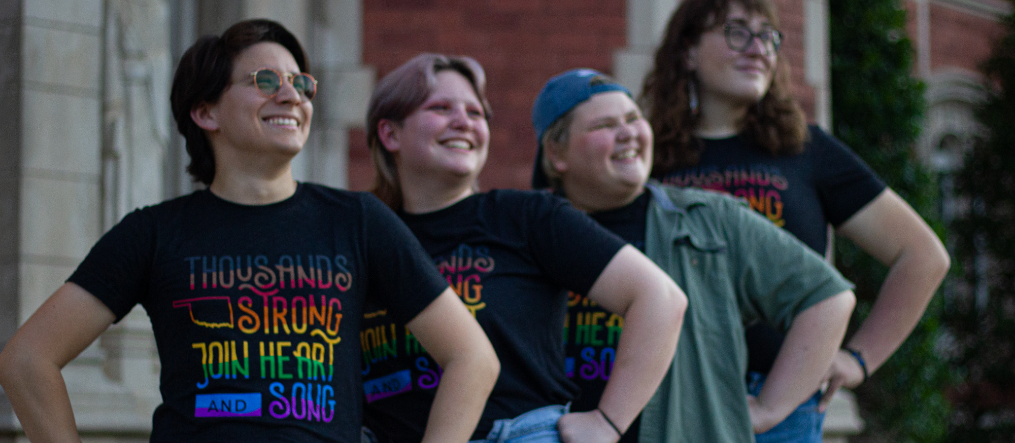 A group of LGBTQIA+ students hands on hip, looking up, smiling, wearing a shirt said "Thousands Strong Join Hearts and Songs"