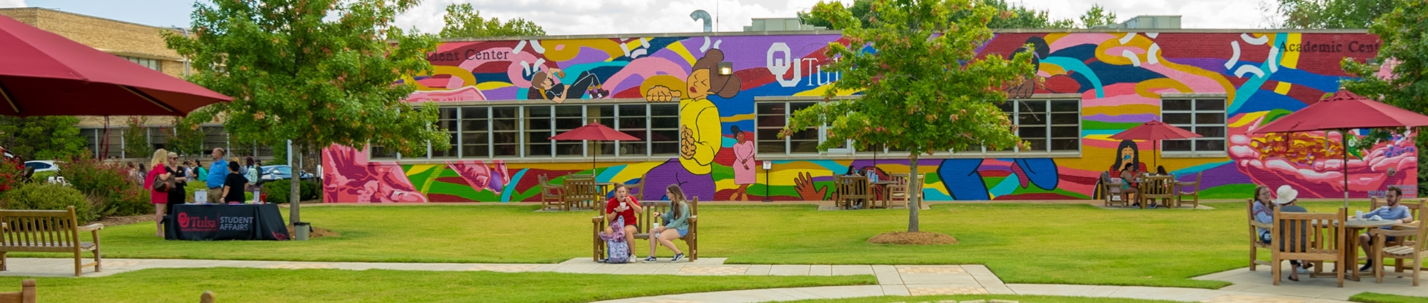 Students sit on benches in a wide green space overlooked by a large, colorful mural in the background