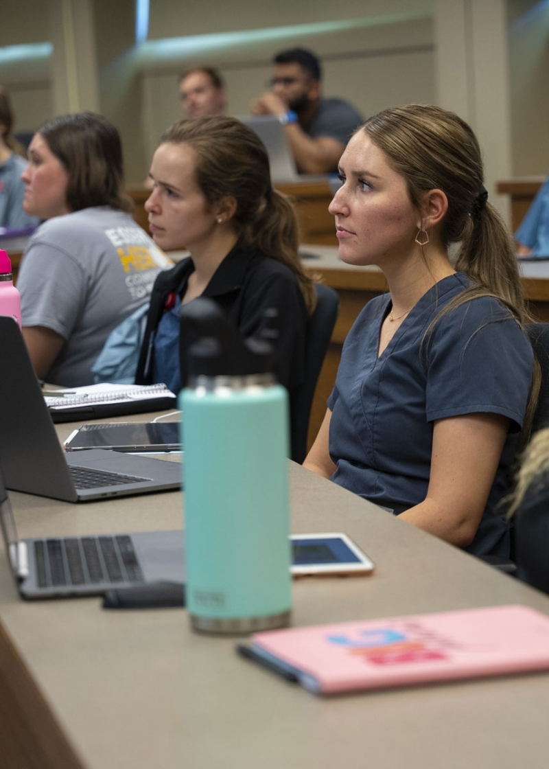A student sits in a classroom listening attentively to a lecture, a lap top and water bottle in front of her