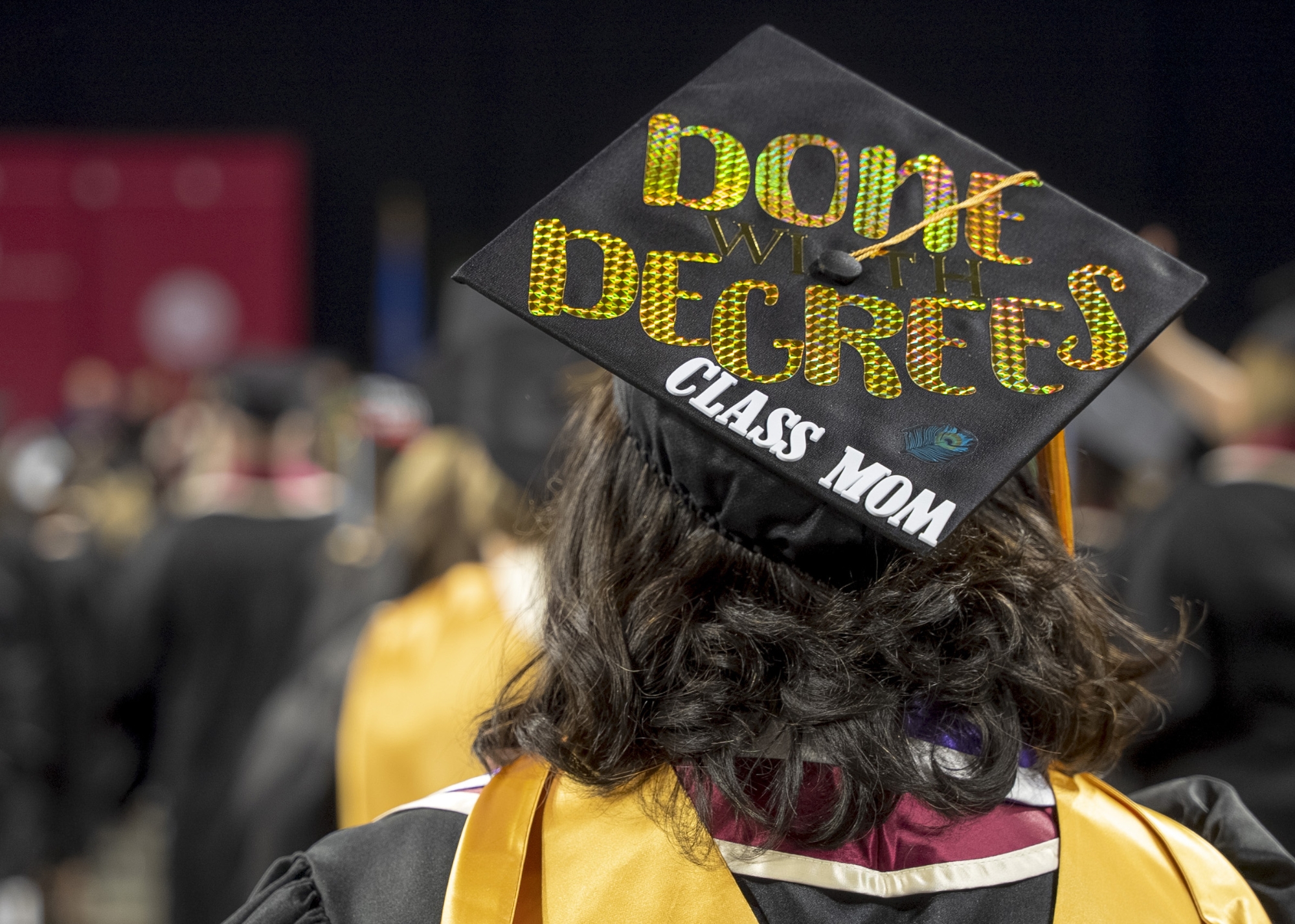 A graduate stands facing the convocation stage. Her mortarboard has been painted with glode letters reading "Done With Degrees" and "Class Mom"