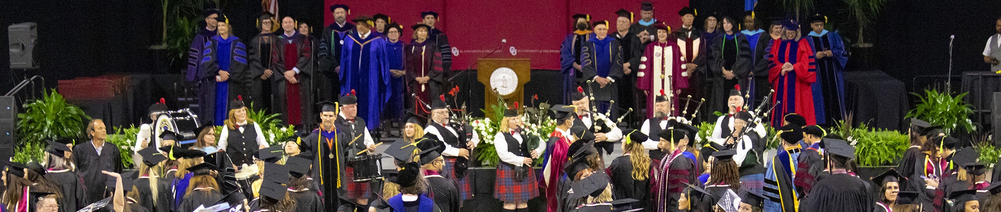 A busy stage is set with faculty in graduation attire of various colors while a Scottish pipe band plays in the foreground. Several students can be seen in black cap and gown