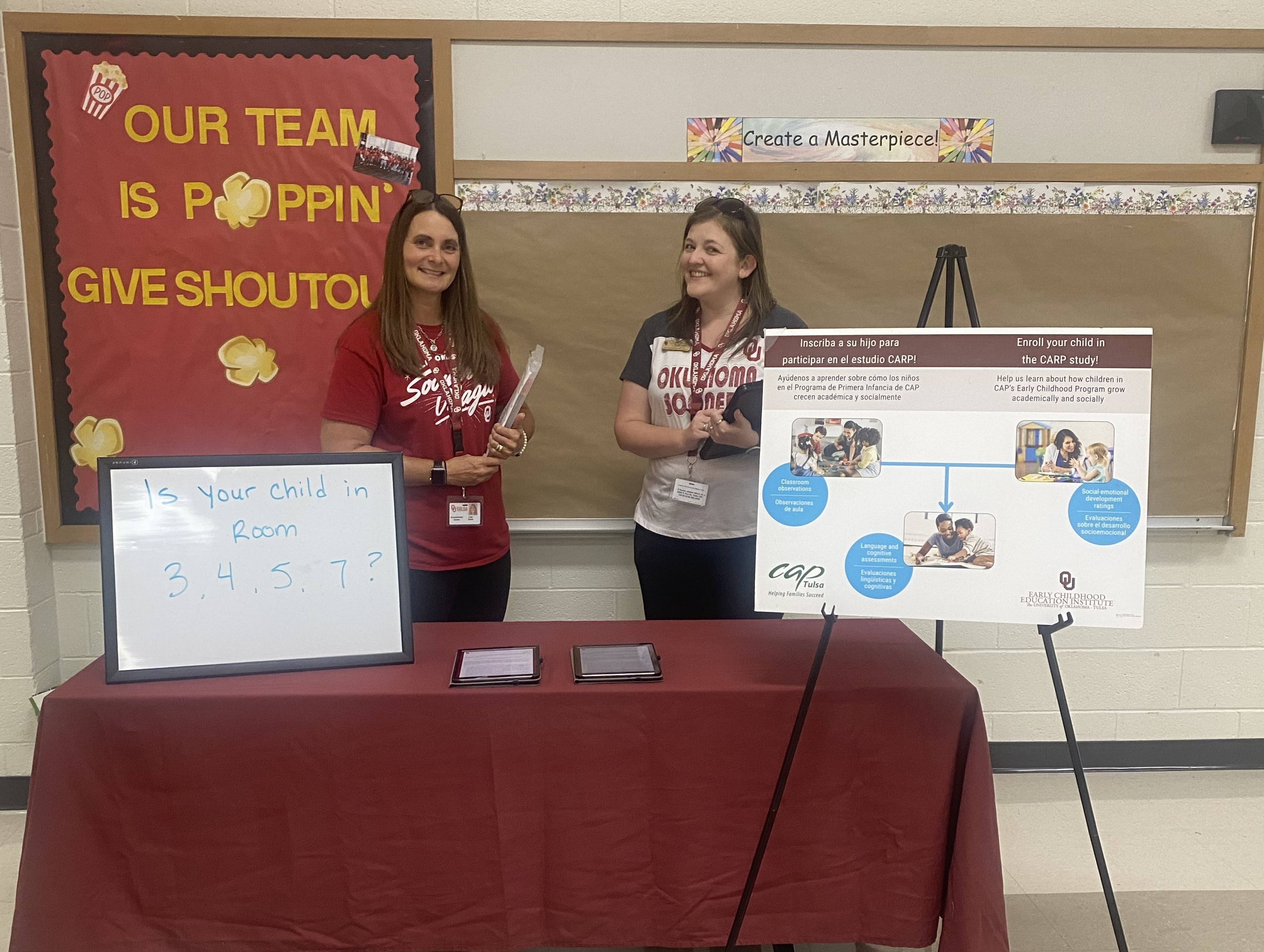 A woman takes notes while a teacher and children work on crafts projects at a table.