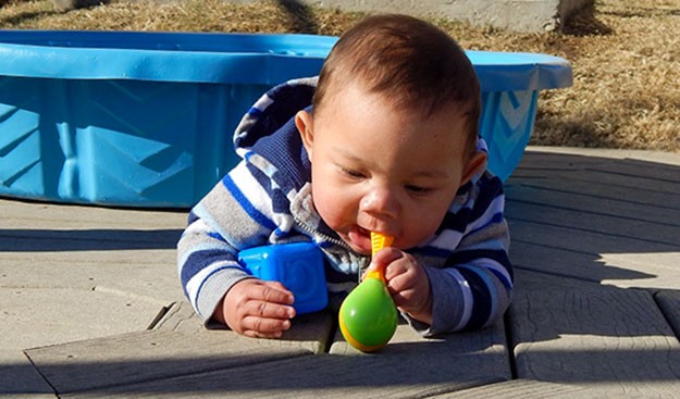 A young boy chews on a plastic rattle outside on a playground.