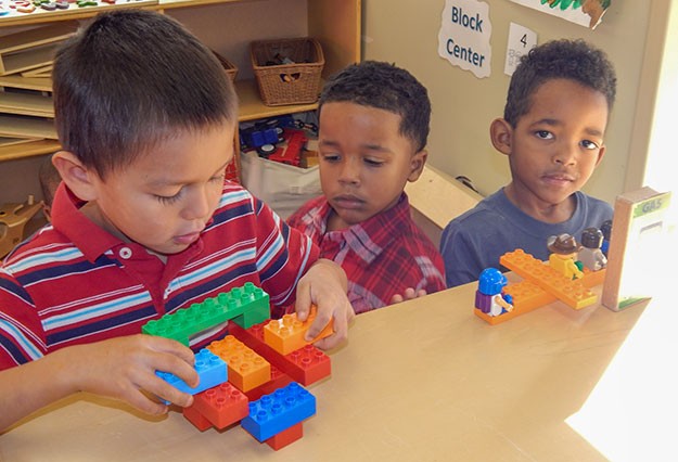 Three boys play with building blocks in a classroom.