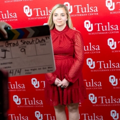 A young woman stands in front of a large red backdrop printed with OU-Tulsa logos while a video slate is held in front of her.