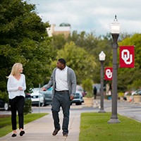 Two students stroll down a sidewalk on a sunny day, flanked by a light post bearing an OU banner