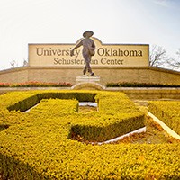 A large OU symbol made of box hedges leads to the feet of a tower statue depicting a man sowing seeds from a bag. A large brink sign behind the statue reads University of Oklahoma Schusterman Center