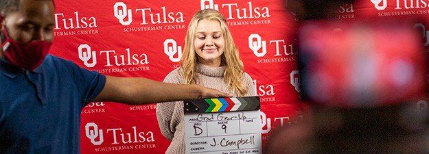 A man holds a video slate in front of a smiling girl who stands in front of a red and white background emblazoned with multiple repeating OU-Tulsa logos.
