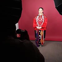 A woman in stunning Native American cultural garb has her photo taken in front of a large, solid crimson background.