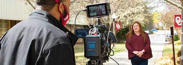 A student with beautiful red hair speaks as a camera flanked by a camera operator records in the foreground