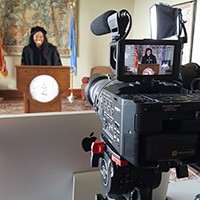 A pretty young woman wearing a graduation cap and gown gives an address standing at a podium as a video camera looks on.