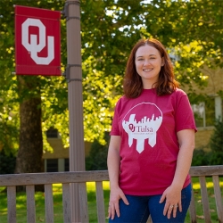A model stands in front of a light post featuring an OU flag. She wears a red t-shirt with a circular white design in the middle that includes a Tulsa skyline and the OU-Tulsa logo
