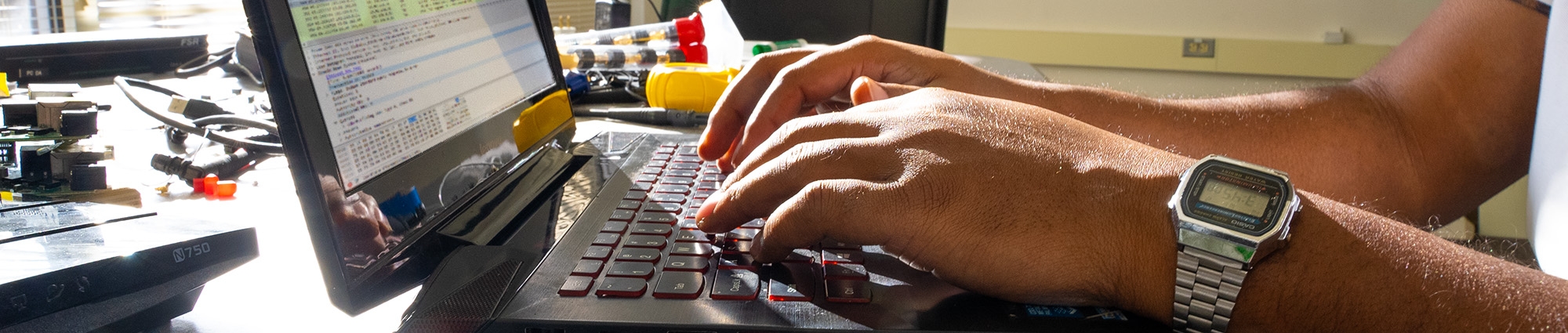 Close up of hands on a laptop keyboard with various computer and engineering related items nearby