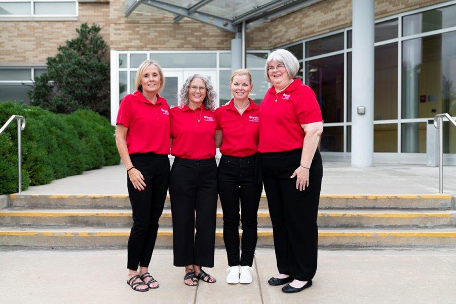 Four staff members wearing red shirts and black vests pose for a group photo outdoors.