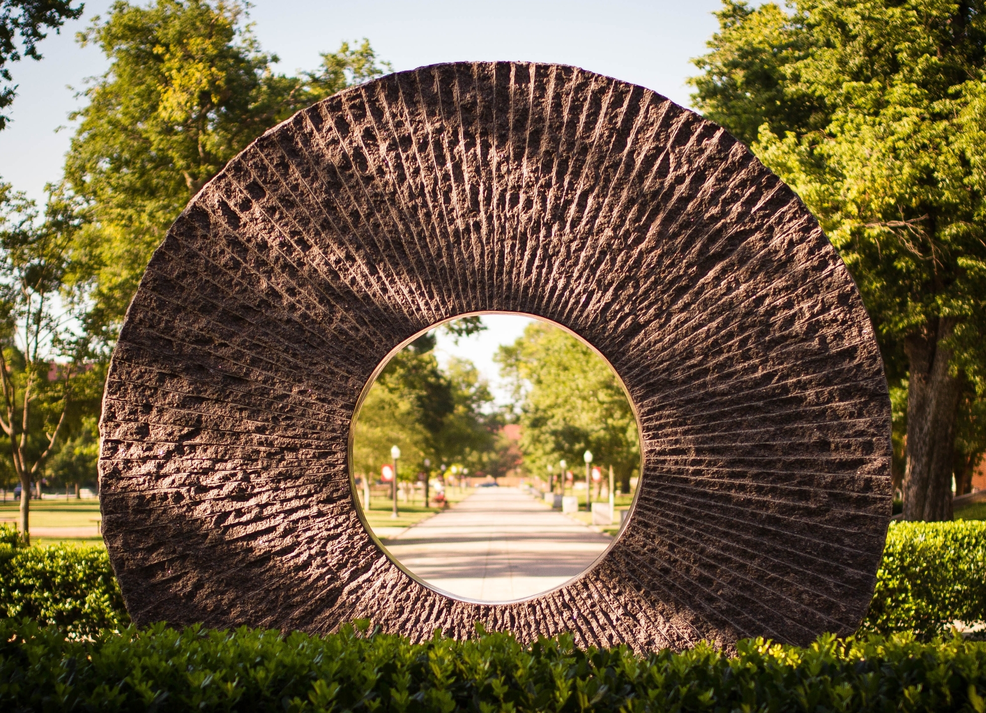 stone disc and bush with trees and sidewalk in the background.