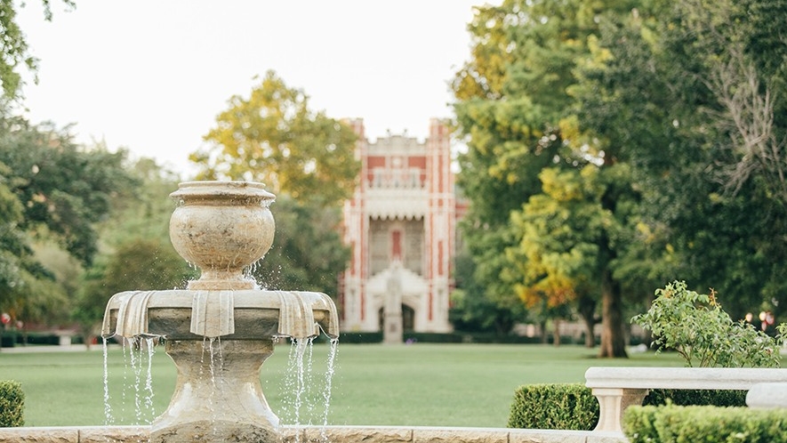 Fountain with a building and trees and grass in the background.